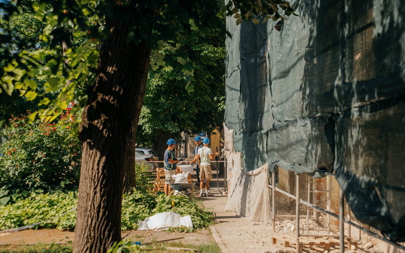Construction workers in blue hard hats stand on a sunny sidewalk beside a building wrapped in tarps, discussing at the site.