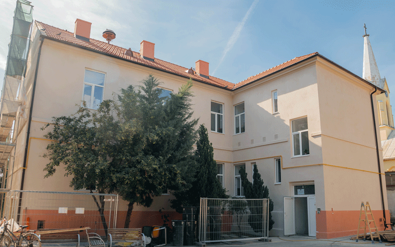 reparatii-scoli4 - Sursa de Vest Beige two-story building with a red tile roof, fenced yard, and green trees in front.