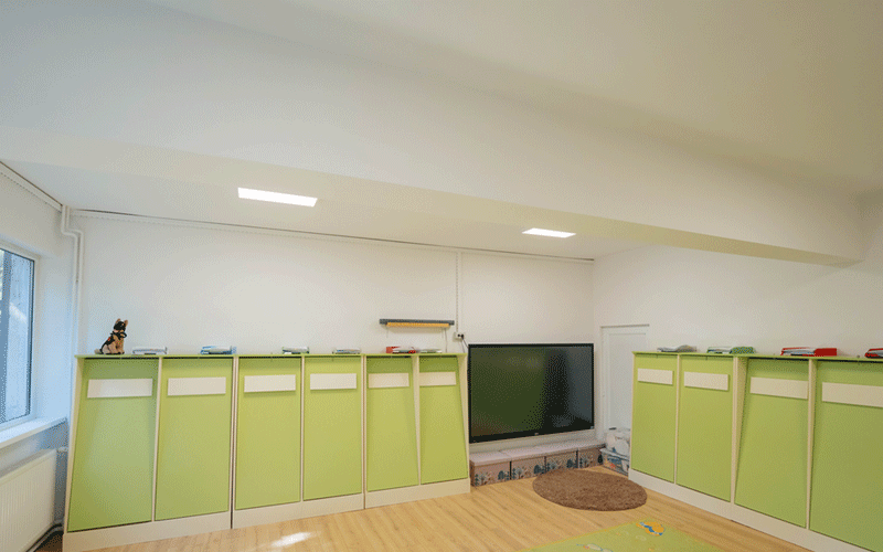 reparatii-scoli5 - Sursa de Vest Bright playroom with lime-green storage cabinets along white walls, a large TV, and a circular brown rug at the center.