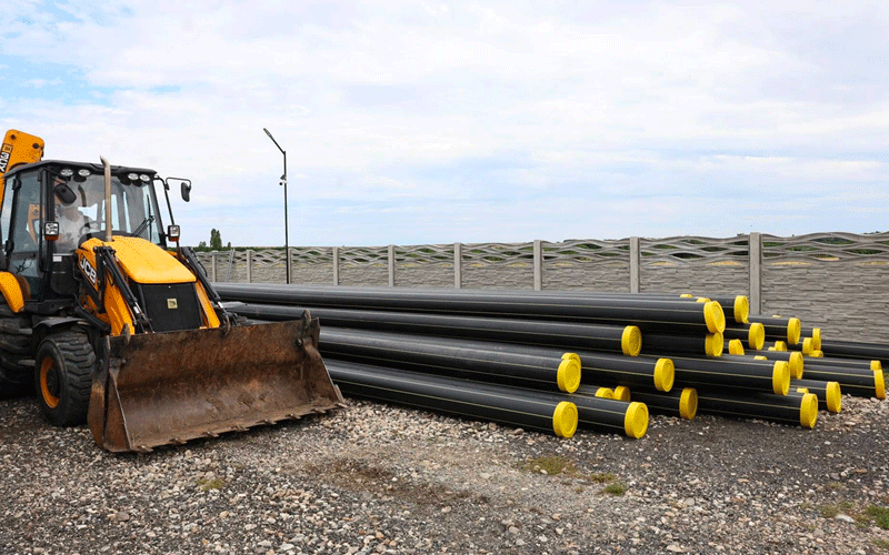 retea-gaz-faget2 - Sursa de Vest Yellow backhoe loader beside a stack of black steel pipes with yellow end caps on a gravel lot, with a fenced, cloudy sky backdrop.