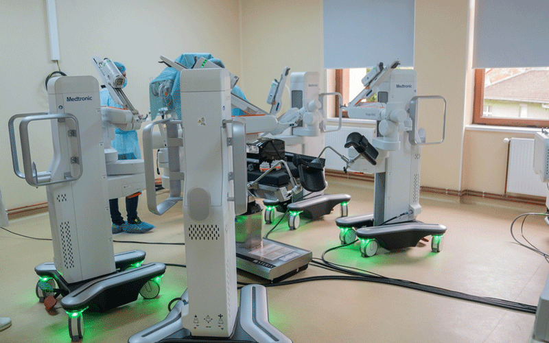 Operating room with multiple robotic surgical arms around a wheeled patient bed and green-lit bases, ready for a procedure.