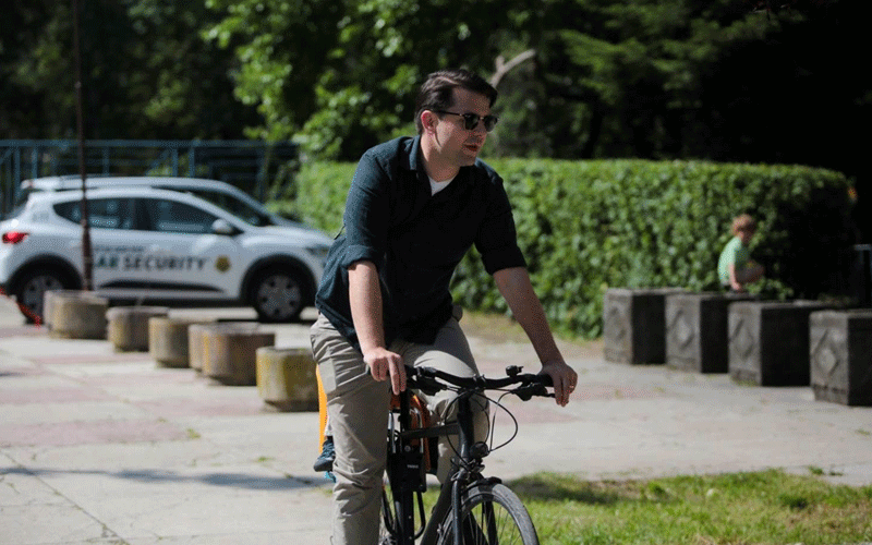 Man wearing sunglasses rides a bicycle on a sunny park path; a security SUV is parked in the background.