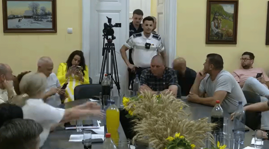 People seated around a conference table in a yellow-walled room; a camera on a tripod stands in the doorway while two officers watch from behind the door.