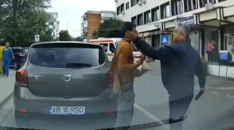 Two men arguing on a city street; a man in a brown shirt is grabbed by the face by another man in a dark jacket, near a gray car with license plate AR 16 RSO behind them.