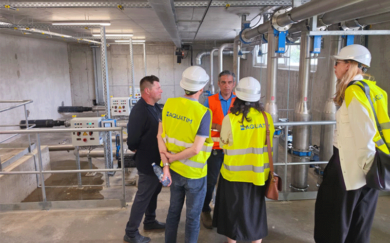 Group of six people in hard hats and high-visibility vests having a discussion in an industrial plant with metal pipes and equipment in the background.