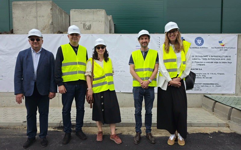 Six adults wearing hard hats and high-visibility vests pose in front of a construction-site banner.