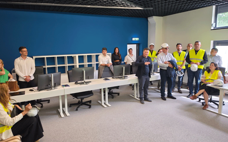 Group of adults in a computer lab, several wearing yellow safety vests, gathered around a long desk with computers for a training session.