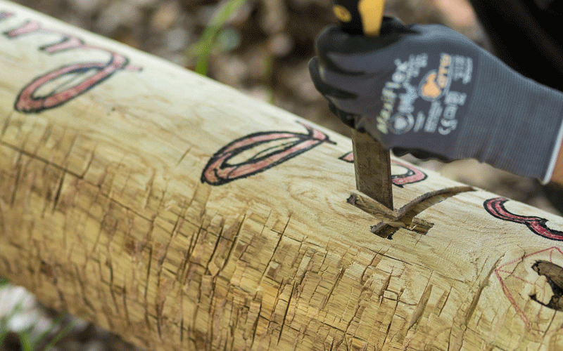 Close-up of a gloved hand chiseling a log, carving decorative shapes into the wood surface.
