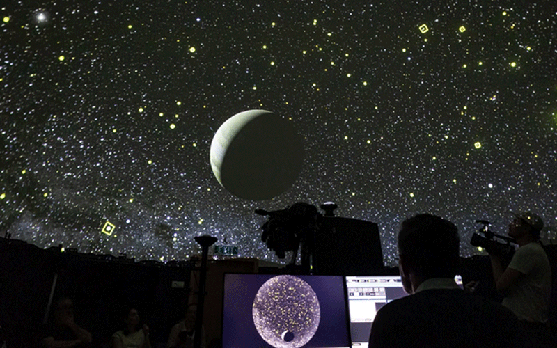 Star-filled night sky with a large Moon projection; silhouettes of visitors and telescope equipment in a dark planetarium.