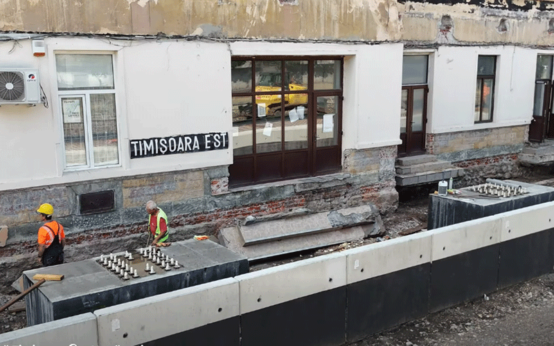 Construction site beside a white building with a 'TIMISOARA EST' sign; two workers in orange vests and helmets near concrete work.