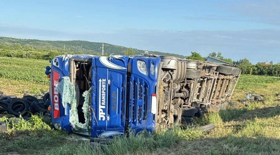 Blue overturned semi-truck on its side in a grassy field, shattered windshield and scattered tires nearby.