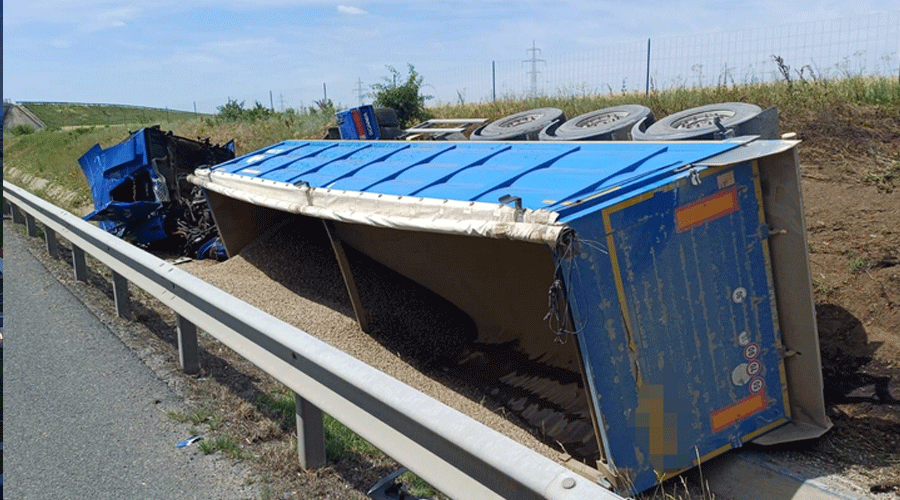 Blue overturned truck trailer spilling gravel onto the highway shoulder beside a guardrail on a sunny day.