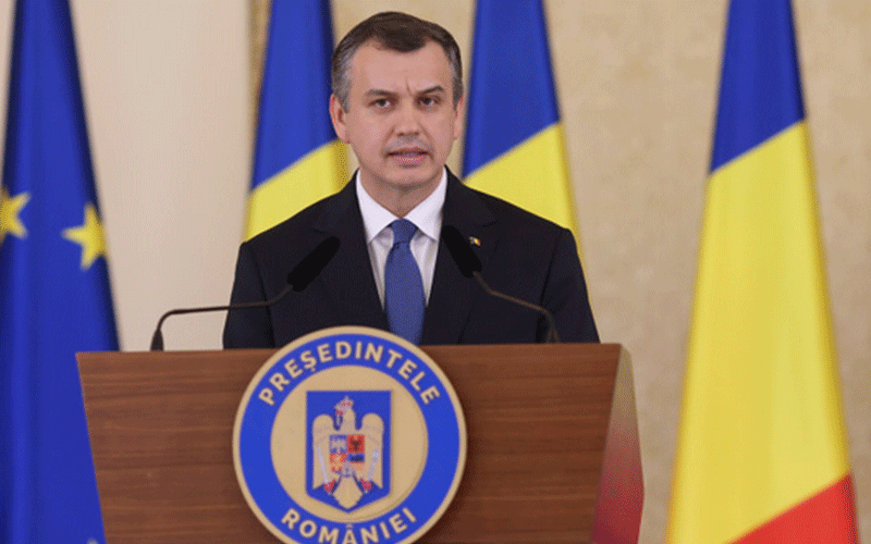 Man in suit giving a formal speech at a wooden podium with the Romanian presidential emblem, flanked by EU and Romanian flags.