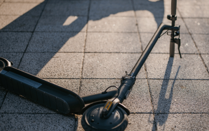 Electric scooter lying on its side on a tiled pavement, with the deck and wheel visible in sunlight