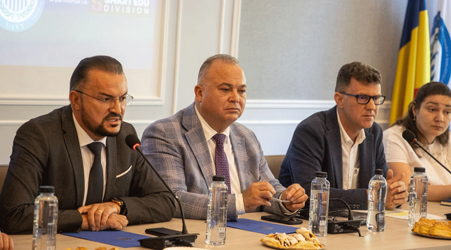 Panel of four professionals at a conference table with microphones, water bottles, and pastries; the man on the left is speaking into a mic.