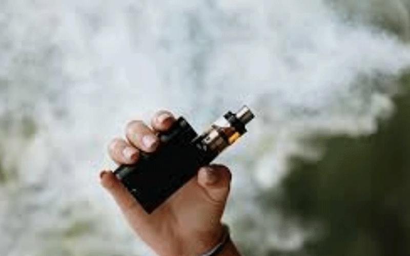 Hand holding a black vaping device (mod) with vapor in the background at outdoors.