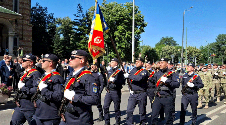Parade of uniformed soldiers marching in formation with a flag bearer, onlookers in background on a sunny day