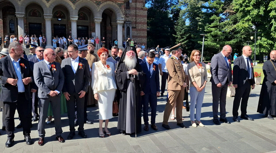 Group of officials and a clergy member standing in a row at an outdoor ceremony in front of a historic arched building.