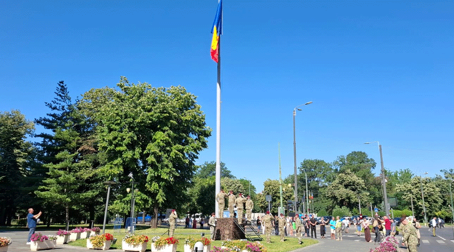 Flagpole with a national flag at a park monument; a crowd of soldiers and civilians gathers on a sunny day.