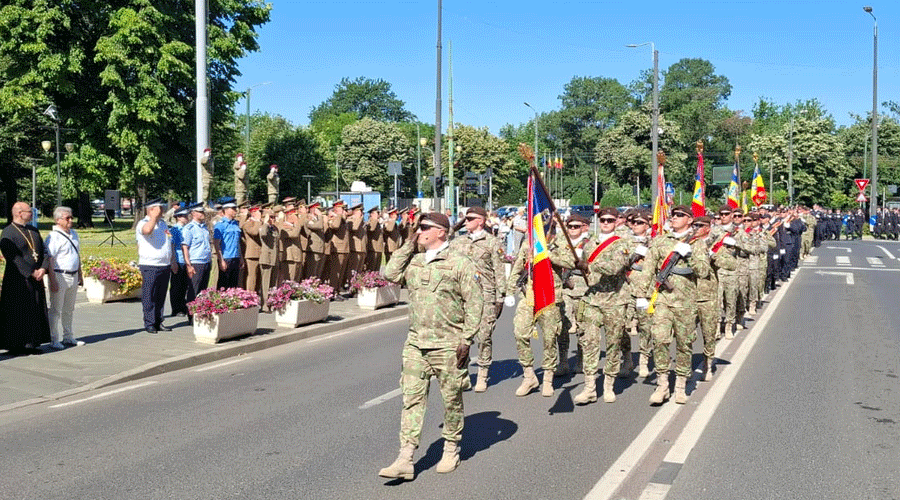 Military parade: soldiers march in formation down a street, carrying flags while spectators watch on a sunny day.