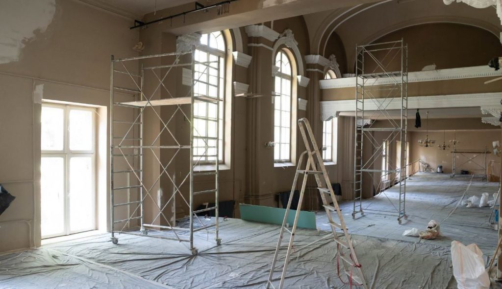 CNB 2 - Sursa de Vest Construction site inside a historic hall with scaffolding, a ladder, and plastic-covered floor. Tall arched windows visible in the background.