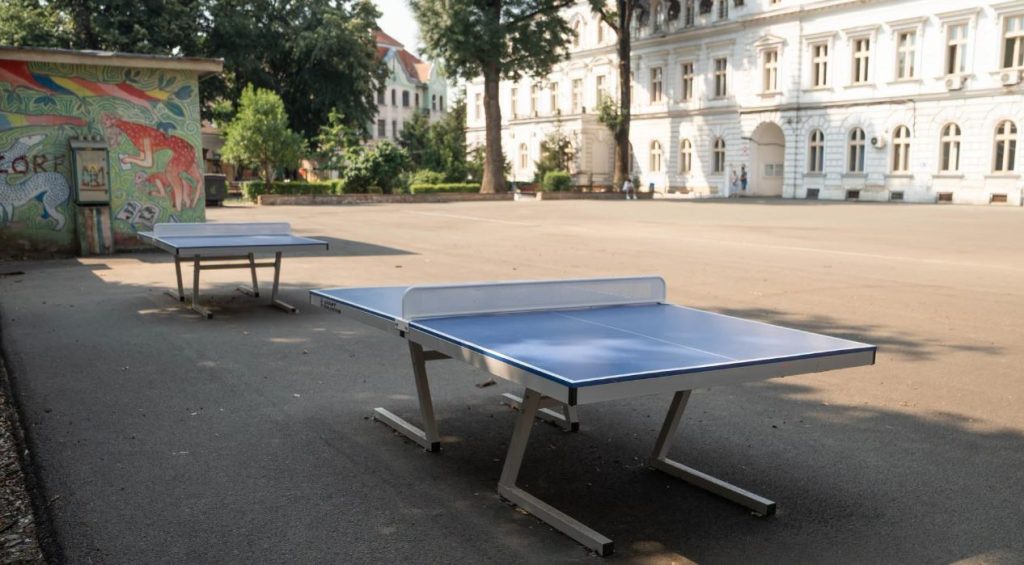 CNB 4 - Sursa de Vest Two blue outdoor ping-pong tables set up on a paved square, with a graffiti wall and a white historic building in the background.