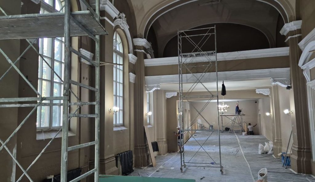 CNB 5 - Sursa de Vest Interior of a grand historic hall under renovation with metal scaffolding, tall arched windows, and workers present on the floor and on scaffolding near ornate molding.