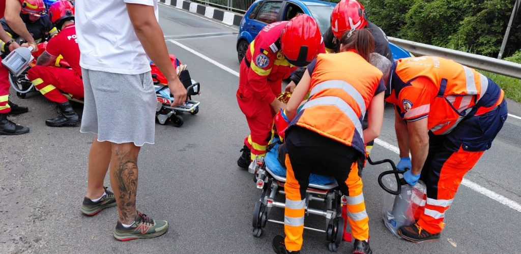 Emergency responders in orange vests move a patient on a wheeled stretcher on a roadway.