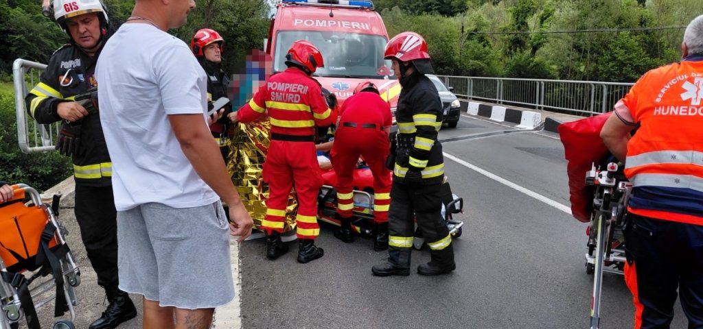 Emergency responders in red and black gear attend to a person on a stretcher beside a red ambulance on a road with a guardrail nearby.