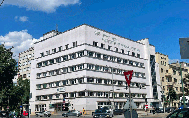 White multi‑story university/campus building with rows of windows under a blue sky, and cars and pedestrians at a busy street in front.