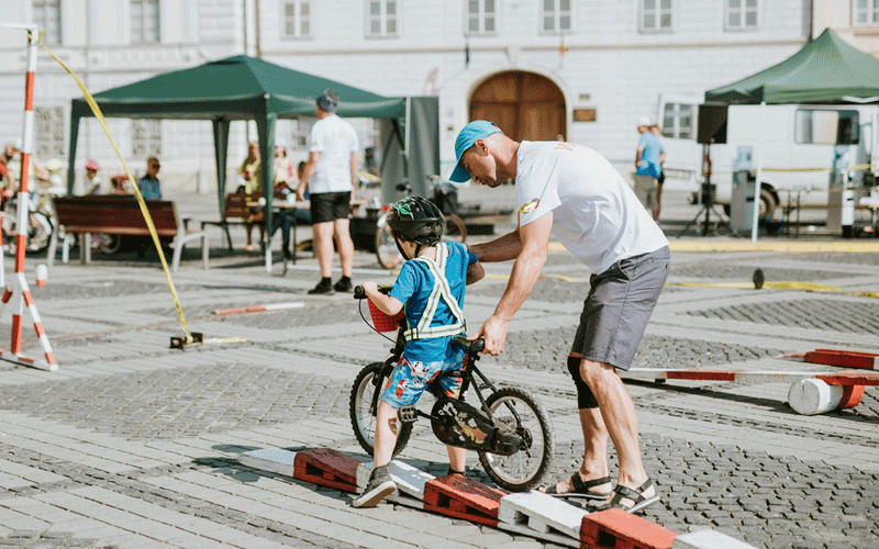 Adult guiding a child on a small bike with training wheels at an outdoor event.