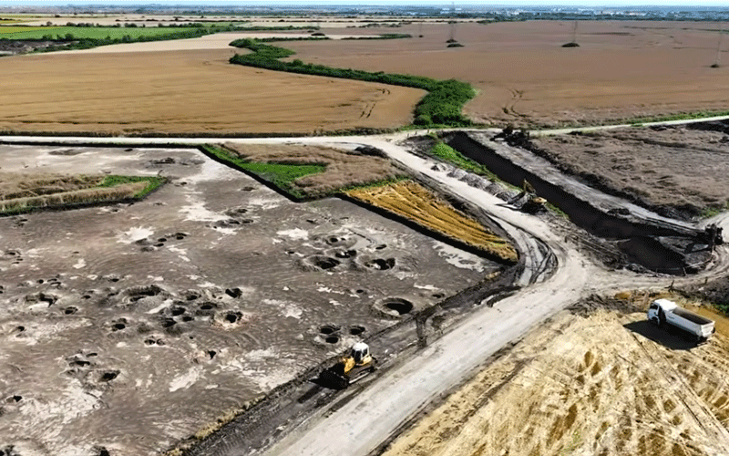 Aerial view of a rural construction site with excavators moving soil along a dirt road, surrounded by fields and green hedges at the edges of farmland.