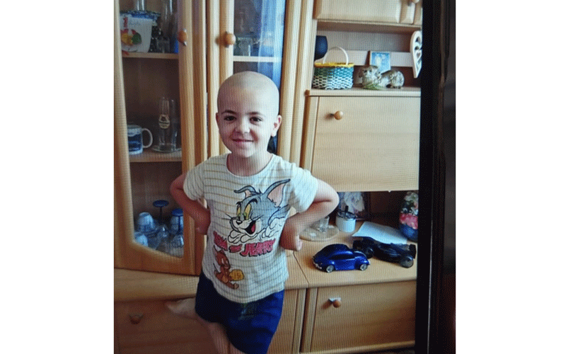 Smiling boy with a shaved head and hands on hips, wearing a Bugs Bunny T‑shirt and blue shorts, standing in a wood cabinet-filled room with toys nearby.