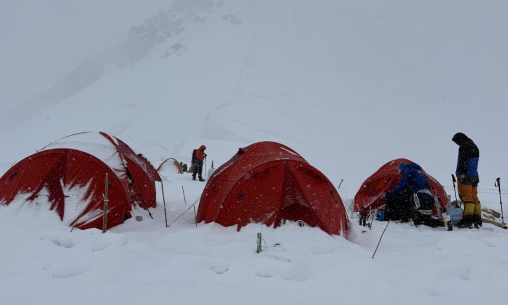 colibasanu 1 - Sursa de Vest Red expedition tents set up in a snowy mountain camp, climbers in heavy snowfall adjusting gear around the tents.