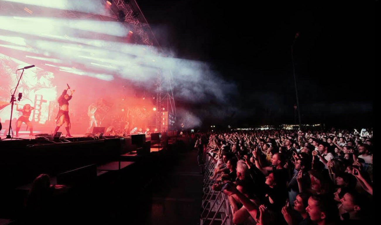 Crowd watches a live concert at night as bright stage lights and smoke illuminate performers on stage ahead.