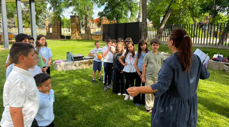 Teacher leads a group of kids outdoors, standing in a semicircle on green grass.