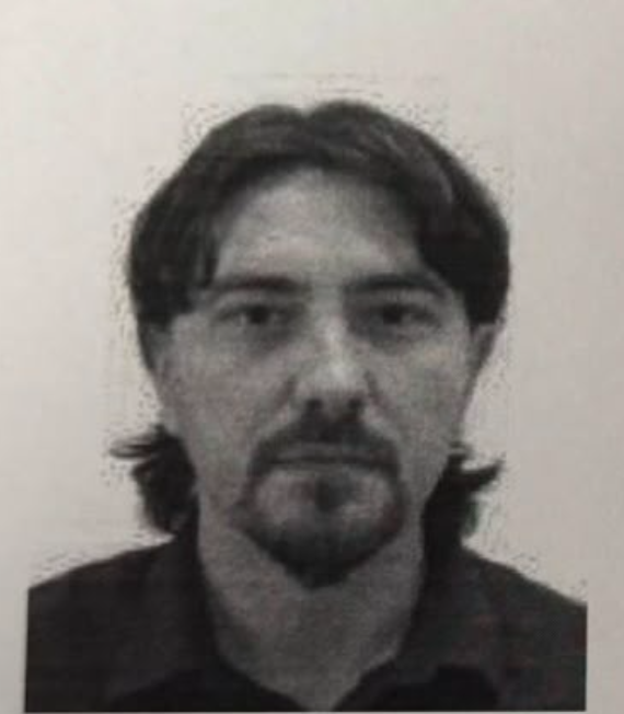 Black-and-white headshot of a man with medium-length dark hair and a goatee, wearing a dark collared shirt, looking at the camera.