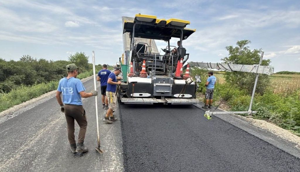 drdp - Sursa de Vest Road construction crew operating a asphalt paver on a rural road, laying new blacktop as cones and workers guide the process.