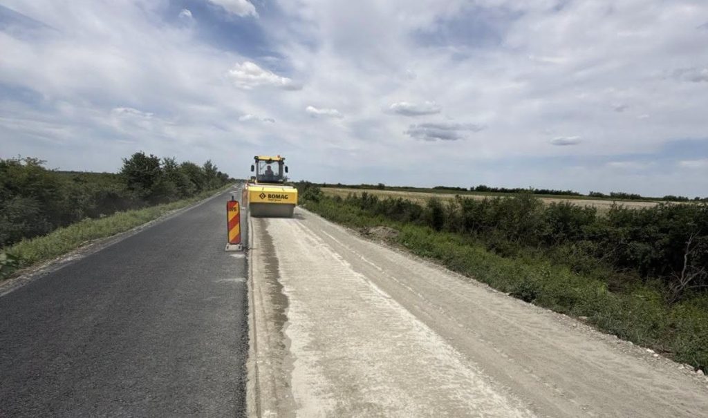 drdp 1 - Sursa de Vest Yellow BOMAG road roller compacting freshly laid asphalt on a rural road; construction barriers nearby as fields and blue sky frame the scene.