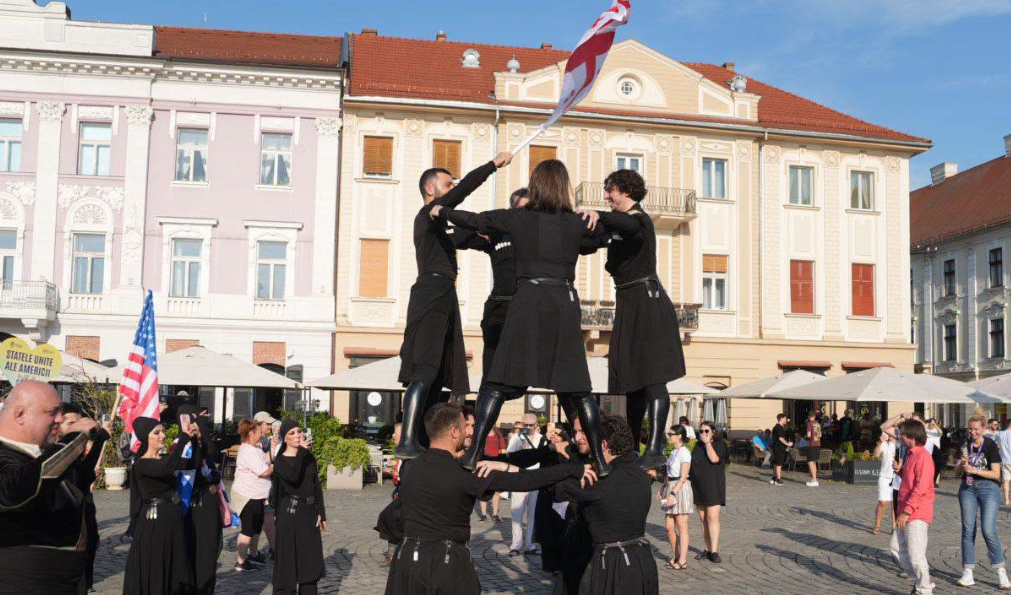 festival inimi2 - Sursa de Vest Group of performers in black outfits form a human pyramid in a town square as spectators watch, with a red-and-white flag overhead.