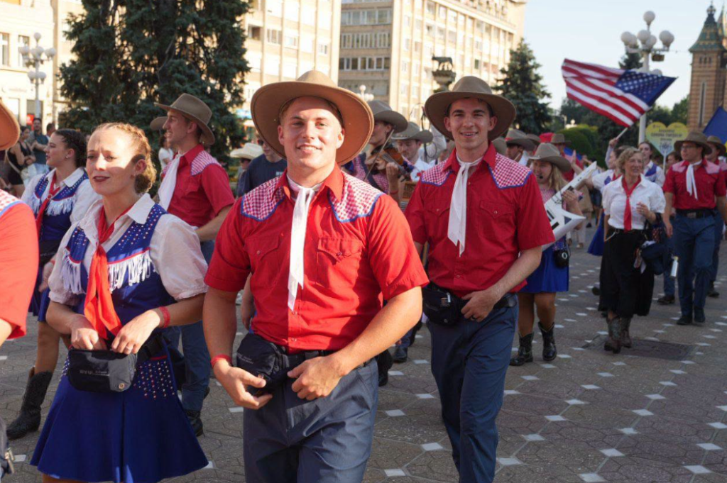 festival inimi3 - Sursa de Vest People in a street parade wearing cowboy outfits, red shirts, hats, and scarves; a lively crowd in the background and an American flag visible.