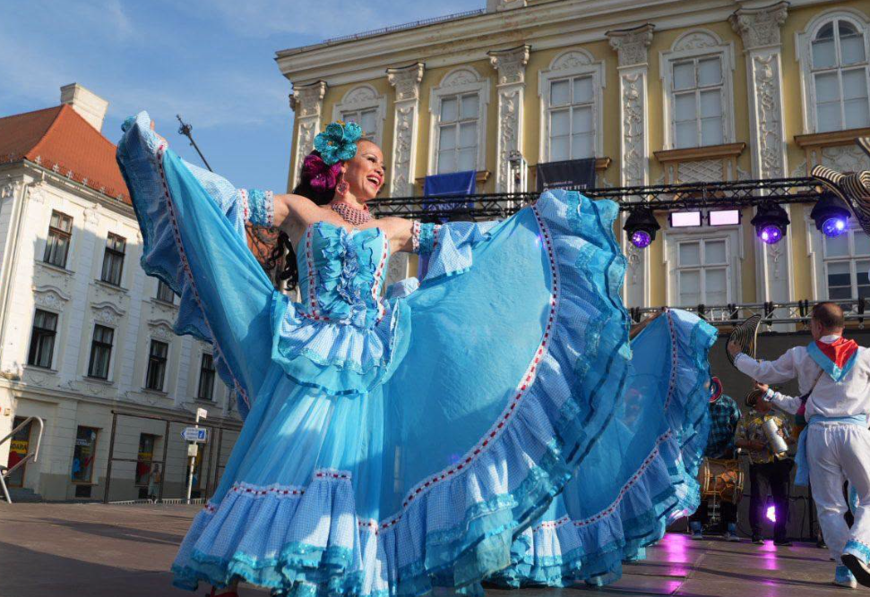 festival inimilor - Sursa de Vest Dancer in a bright blue flowing dress twirls on an outdoor stage in front of historic buildings.