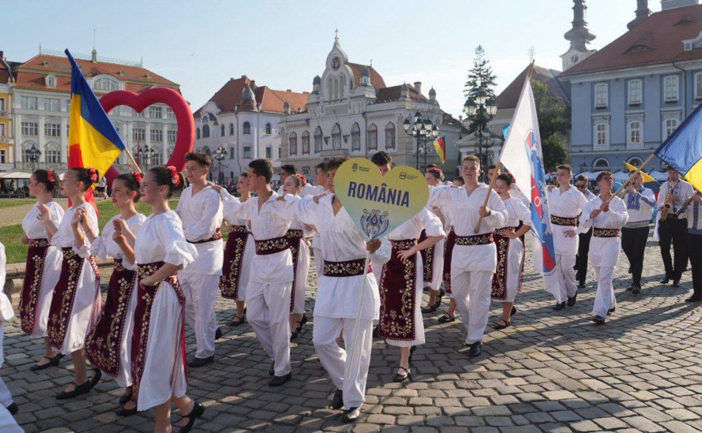 festivalul inimilor - Sursa de Vest People in white Romanian folk costumes march in a parade on a cobblestone square, holding a heart-shaped ROMANIA sign and flags.