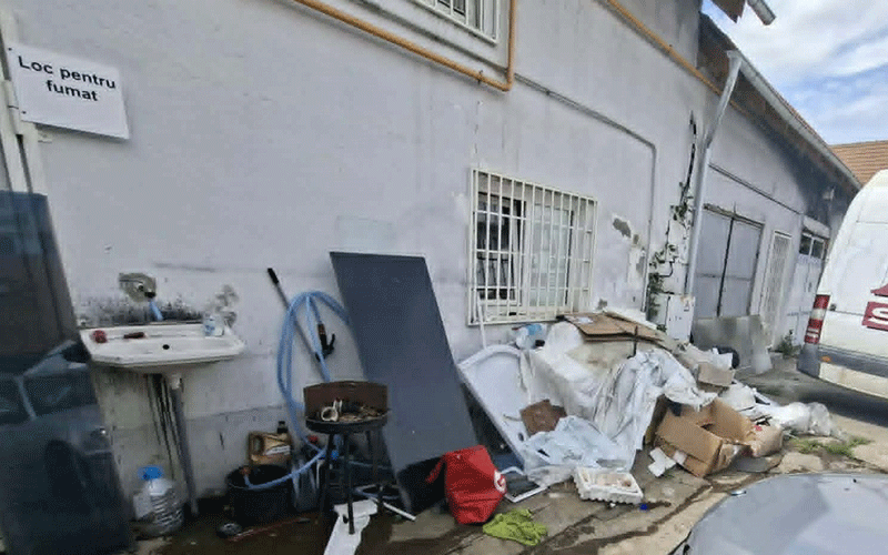 Exterior alley next to a pale building with a barred window, cluttered with garbage, boxes, mattresses, and discarded items along the wall; a van is parked nearby.