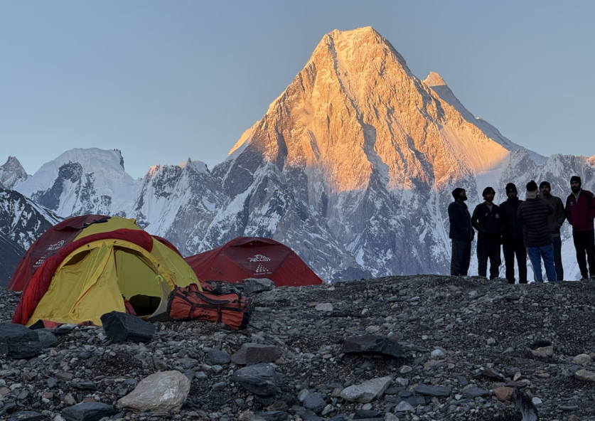 horia colibasanu - Sursa de Vest Group of hikers standing together on a rocky tundra near red and yellow tents with towering snow-capped mountains at sunrise behind them.