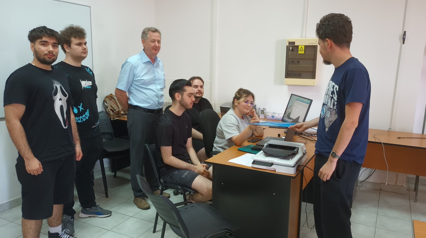Group of students in a classroom gathered around a desk with a laptop and printer while a man explains something. Note: concise, informative.