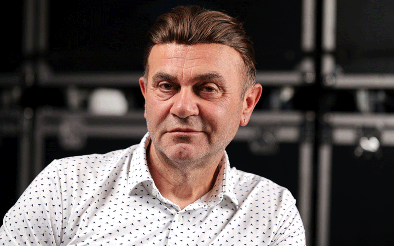 Portrait of a middle-aged man in a white patterned shirt facing the camera, with a blurred backstage setup in the background.