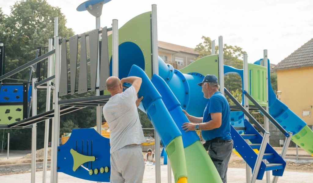 loc de joaca 2 - Sursa de Vest Two men assembling a colorful outdoor playground with blue and green tubes and slides on a sunny day.