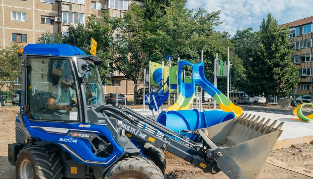 loc de joaca 3 - Sursa de Vest Blue compact front loader with bucket at a construction site beside a colorful children's playground in a residential area.