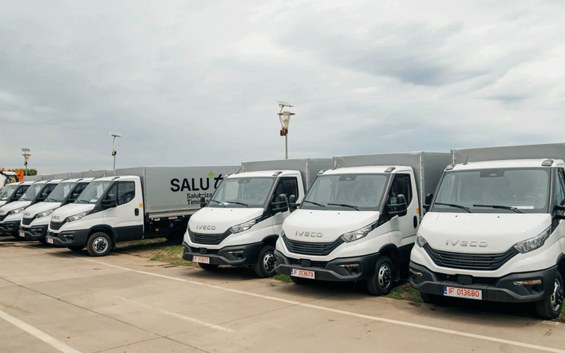 Line of white Iveco delivery trucks parked in an outdoor lot with a long trailer in the background and an overcast sky.